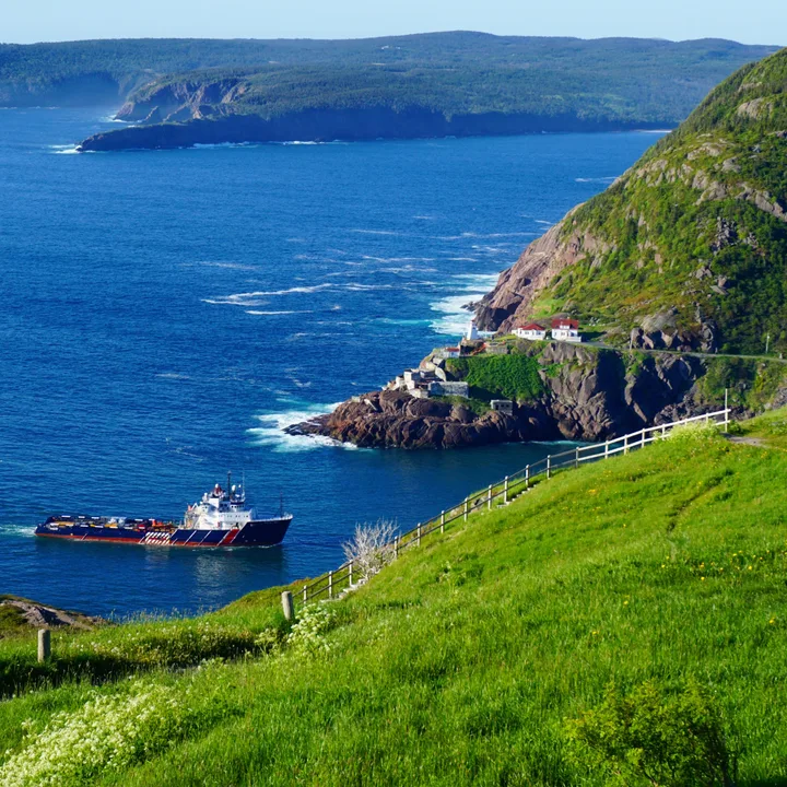 A red and white supply vessel entering a narrow harbour channel below steep green headlands, with a white lighthouse on the rocks.