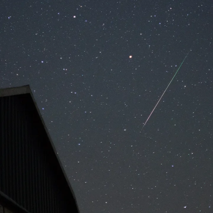 A meteor with a green and white tail streaking across a dense field of stars, above the dark silhouette of a barn roof.