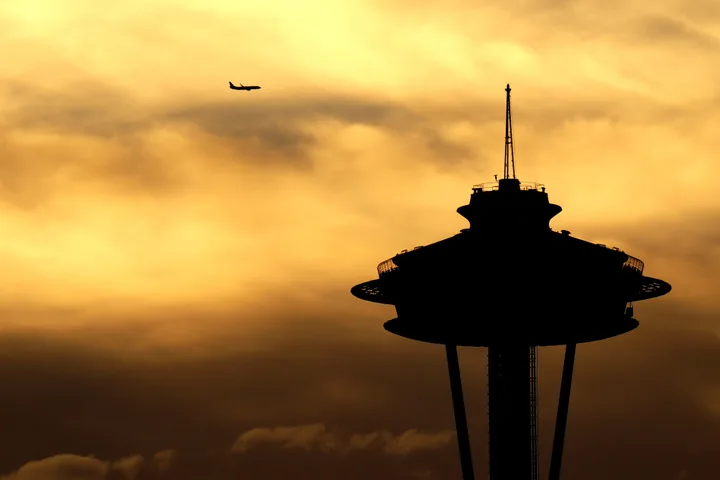 The Space Needle in full silhouette against a burning orange sunset sky, with a distant airliner passing at upper left.