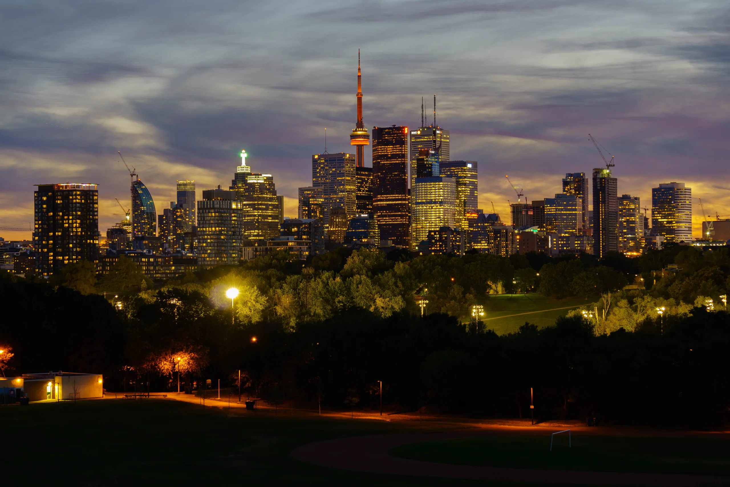The Toronto skyline at night, the CN Tower lit orange above the downtown towers and a last band of sunset burning low behind them, with a floodlit park in the dark foreground.